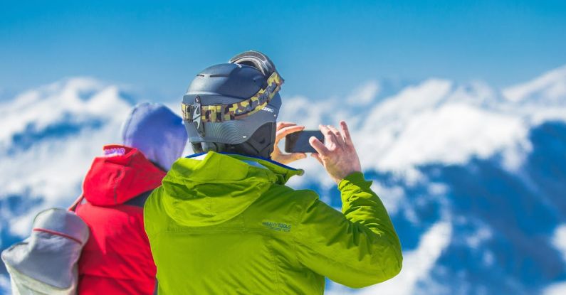 Winter Gear - Man in Green Jacket and Gray Helmet Holding Phone Standing Next to Person in Red and Black Jacket