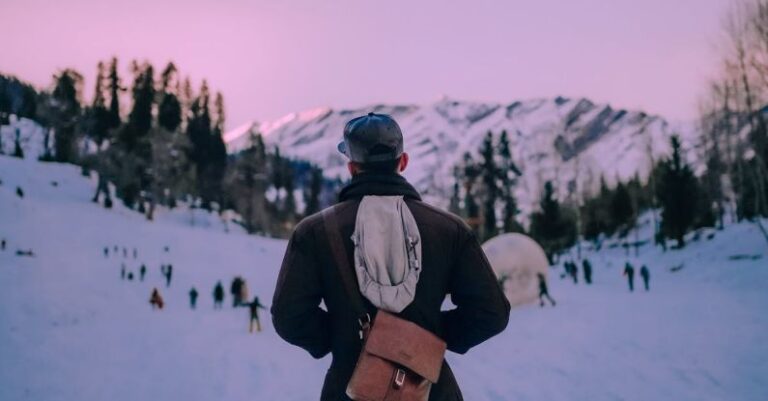 Ski Trip - Back view anonymous male traveler in warm clothes standing with hands in pockets on snowy mountain slope in modern ski resort at twilight